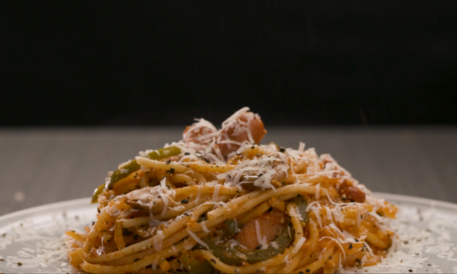 A close-up of spaghetti mixed with vegetables and topped with grated cheese on a white plate with a dark background.